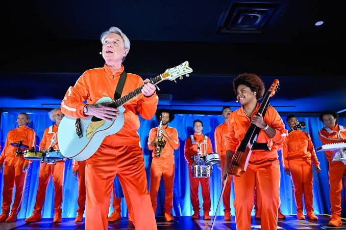 Live performance with a band in matching orange outfits on a blue-lit stage